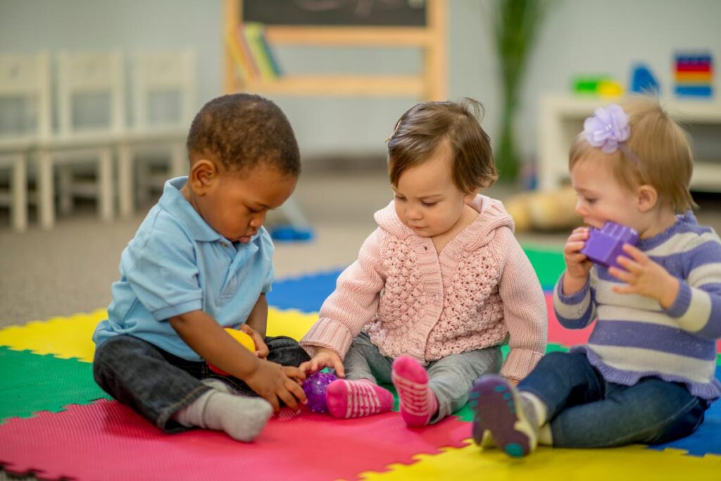 Three toddlers sit on a colorful foam mat in a classroom, playing with toys. Two children are focused on stacking colorful blocks, while one child holds a purple block to their mouth. Shelves and a chalkboard are visible in the background.