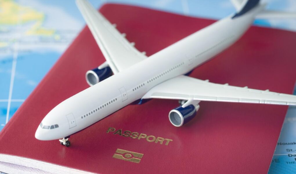 A toy airplane rests on a closed red passport with a gold emblem and the word "PASSPORT" on it, placed on a colorful world map showing parts of continents and oceans, symbolizing travel and international journeys.