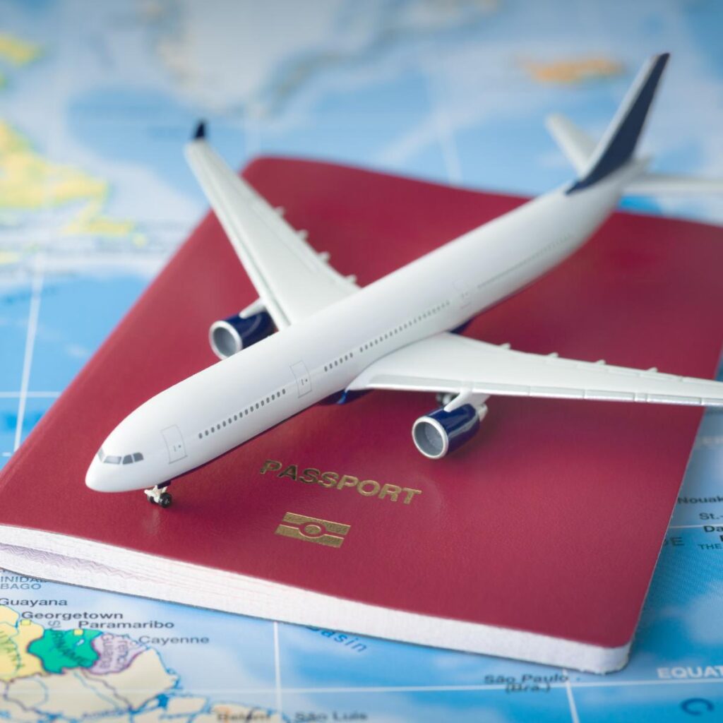 A toy airplane rests on a closed red passport with a gold emblem and the word "PASSPORT" on it, placed on a colorful world map showing parts of continents and oceans, symbolizing travel and international journeys.