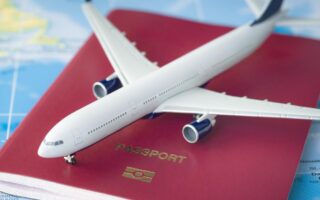 A toy airplane rests on a closed red passport with a gold emblem and the word "PASSPORT" on it, placed on a colorful world map showing parts of continents and oceans, symbolizing travel and international journeys.