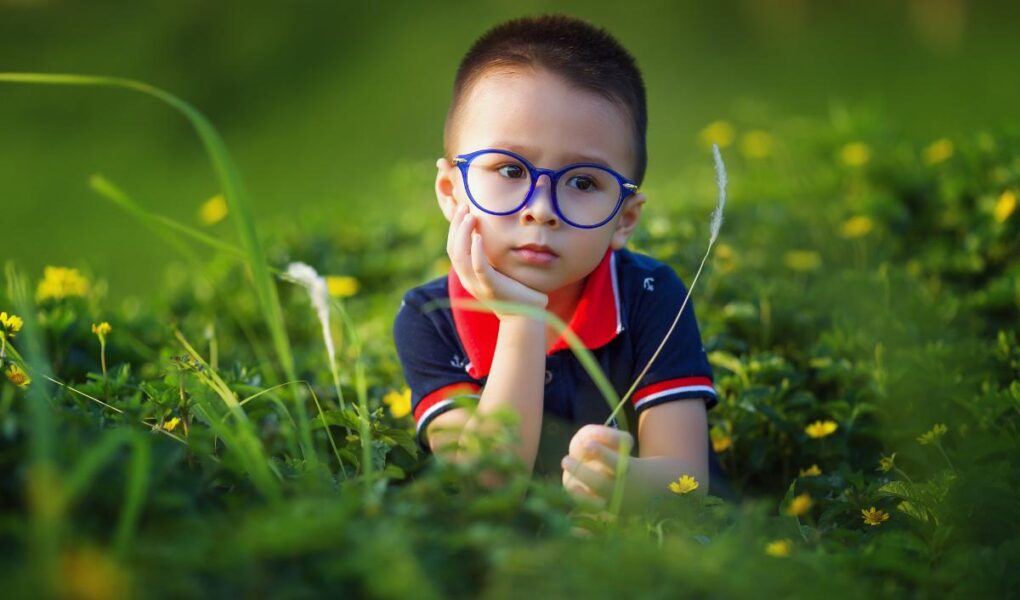 A young child with short hair and blue glasses sits in a field of green grass and yellow flowers, resting their chin on one hand and looking thoughtful. The child wears a navy blue shirt with a red collar. The background is softly blurred.