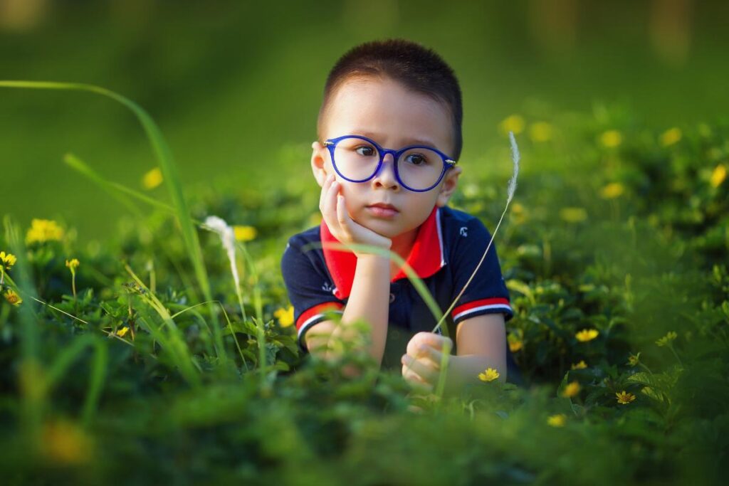 A young child with short hair and blue glasses sits in a field of green grass and yellow flowers, resting their chin on one hand and looking thoughtful. The child wears a navy blue shirt with a red collar. The background is softly blurred.