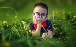 A young child with short hair and blue glasses sits in a field of green grass and yellow flowers, resting their chin on one hand and looking thoughtful. The child wears a navy blue shirt with a red collar. The background is softly blurred.