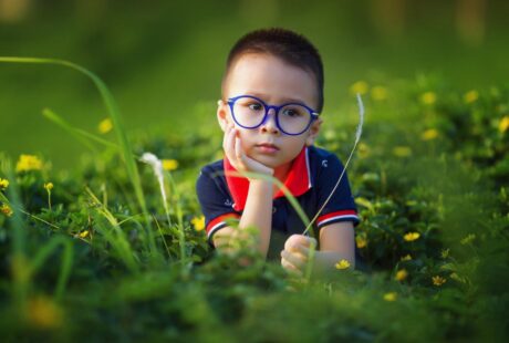 A young child with short hair and blue glasses sits in a field of green grass and yellow flowers, resting their chin on one hand and looking thoughtful. The child wears a navy blue shirt with a red collar. The background is softly blurred.