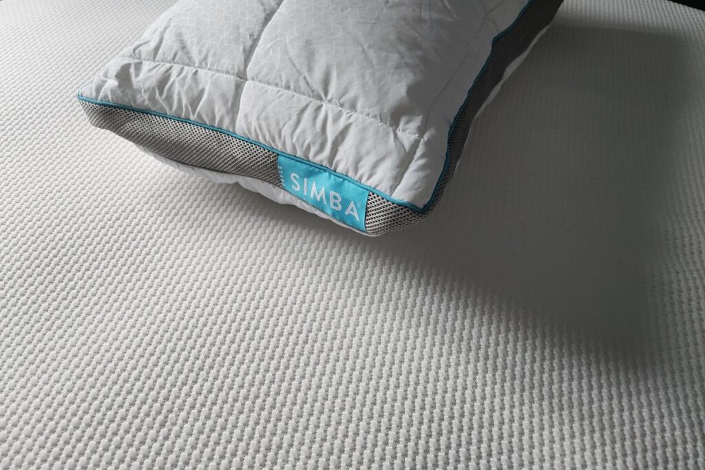 A white pillow with a blue edge and a "SIMBA" label rests on a textured simba hybrid mattress. The subtle waffle-like pattern catches sunlight, casting soft shadows and highlighting the fabric’s surface. Only part of the pillow is visible.