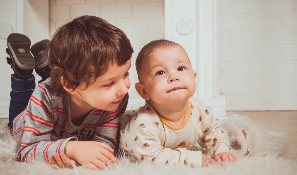 Two young children lie on a fluffy white rug indoors. The older child, in a striped shirt, smiles while leaning close to the baby, who wears a light-colored onesie with patterns and gazes upward. They appear relaxed and content in a cozy setting.