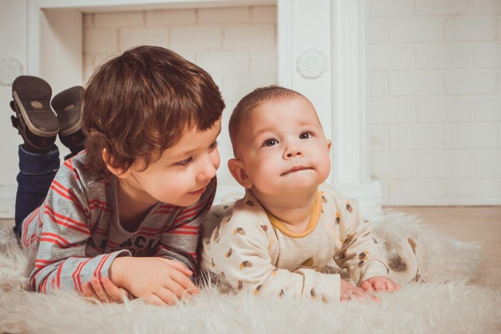 Two young children lie on a fluffy white rug indoors. The older child, in a striped shirt, smiles while leaning close to the baby, who wears a light-colored onesie with patterns and gazes upward. They appear relaxed and content in a cozy setting.