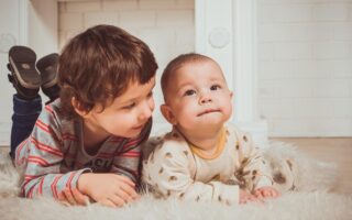 Two young children lie on a fluffy white rug indoors. The older child, in a striped shirt, smiles while leaning close to the baby, who wears a light-colored onesie with patterns and gazes upward. They appear relaxed and content in a cozy setting.