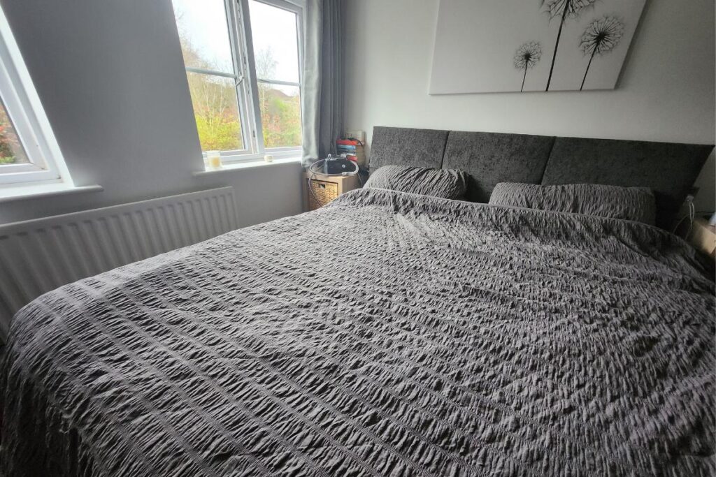 A neatly made bed with a textured gray bedspread and matching pillows, featuring a Simba Hybrid mattress, is against a white wall. Above the bed hangs black-and-white dandelion artwork. Sunlight filters through two windows on the left, beside a nightstand.