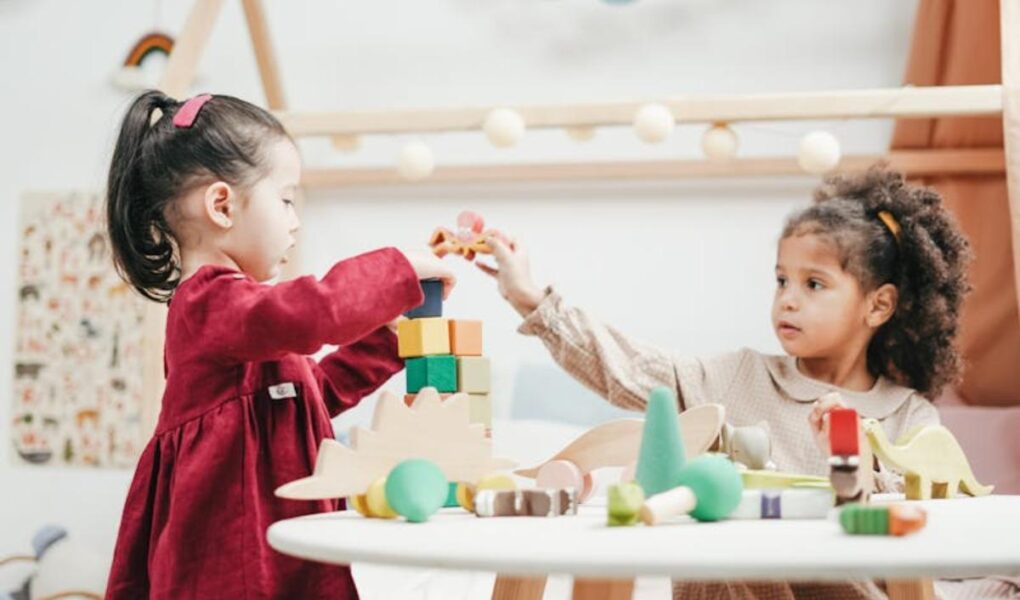Two young girls play together at a white table, building a tower with colorful wooden blocks and toy dinosaurs in a bright, cozy room decorated with string lights and playful accents. One girl wears a red dress, the other a beige dress.