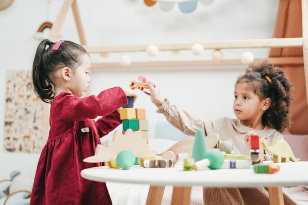 Two young girls play together at a white table, building a tower with colorful wooden blocks and toy dinosaurs in a bright, cozy room decorated with string lights and playful accents. One girl wears a red dress, the other a beige dress.