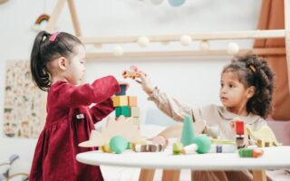Two young girls play together at a white table, building a tower with colorful wooden blocks and toy dinosaurs in a bright, cozy room decorated with string lights and playful accents. One girl wears a red dress, the other a beige dress.