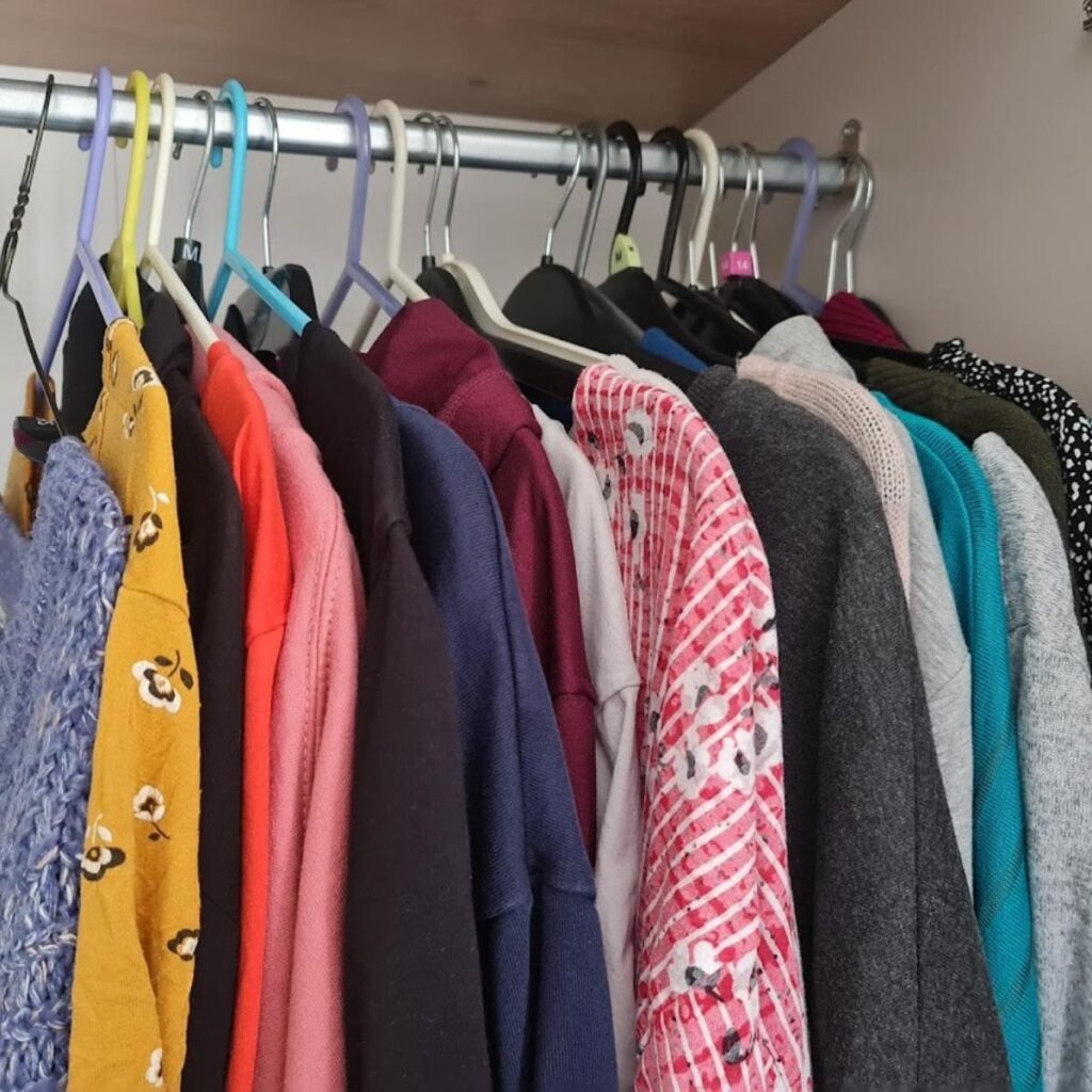 A row of various sweaters, cardigans, and tops in different colors and patterns hang on a metal rod in a closet. The garments are on plastic and metal hangers and include yellow, pink, purple, gray, striped, and floral pieces.