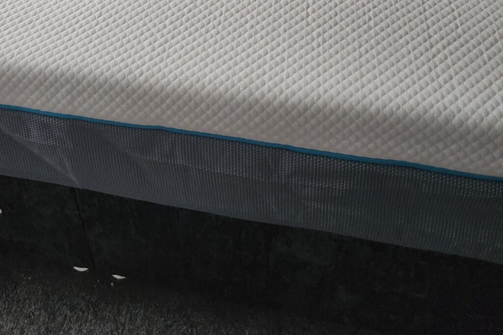 A close-up of a Simba Hybrid mattress with a white, textured quilted surface and a dark blue fabric border. The mattress sits on a black bed base, and the floor below is covered in dark gray carpet.