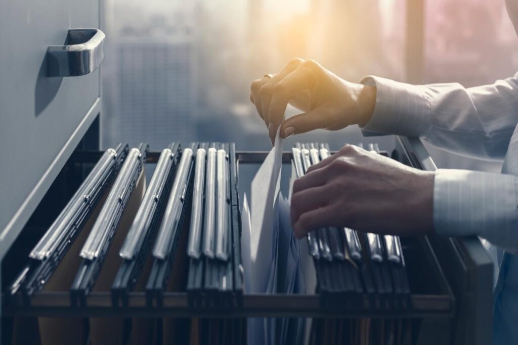 A person sorts through hanging file folders in an open filing cabinet drawer, with city buildings visible through a window in the background. Warm sunlight streams into the office, illuminating the person’s hands and the documents.