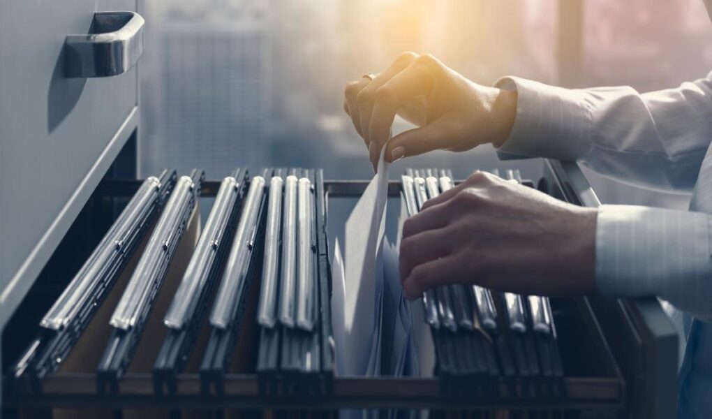 A person’s hands are sorting through hanging file folders in an open filing cabinet drawer. Sunlight filters through a large window in the workspace, revealing a cityscape with tall, blurry buildings—suggesting office organization or document management.