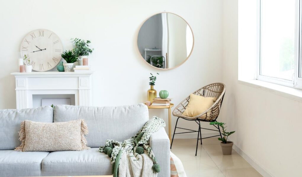 Bright, minimalist living room with a light gray sofa, beige textured pillow, green throw blanket, round mirror, wicker chair with yellow cushion, white mantel with plants and a clock, large window, tiled floor, and decor accents like magazines and vases.