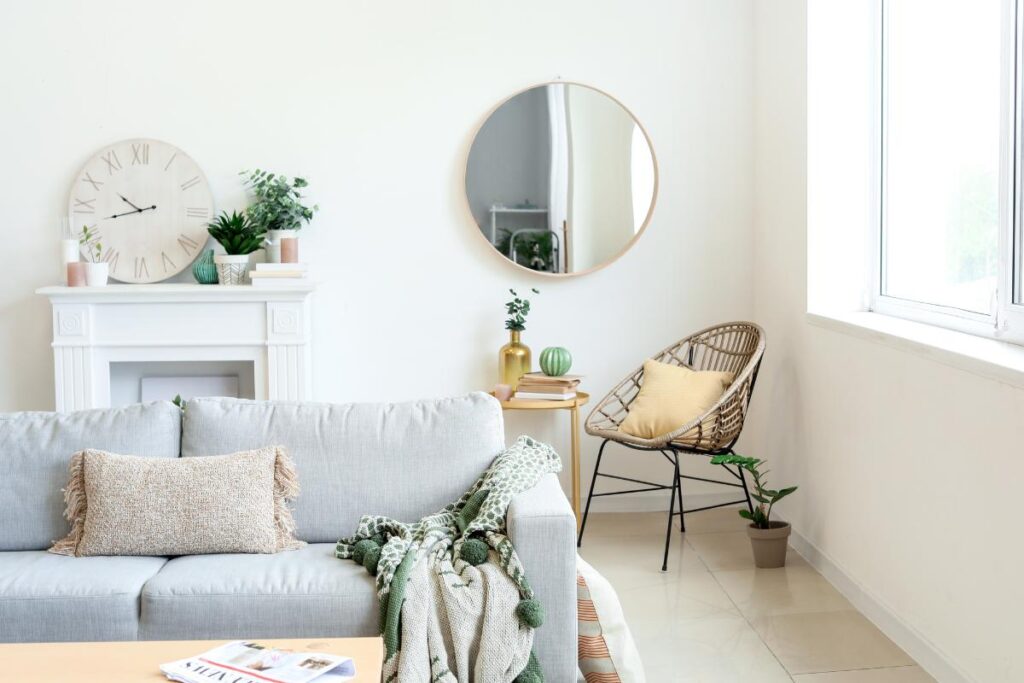 Bright, minimalist living room with a light gray sofa, beige textured pillow, green throw blanket, round mirror, wicker chair with yellow cushion, white mantel with plants and a clock, large window, tiled floor, and decor accents like magazines and vases.