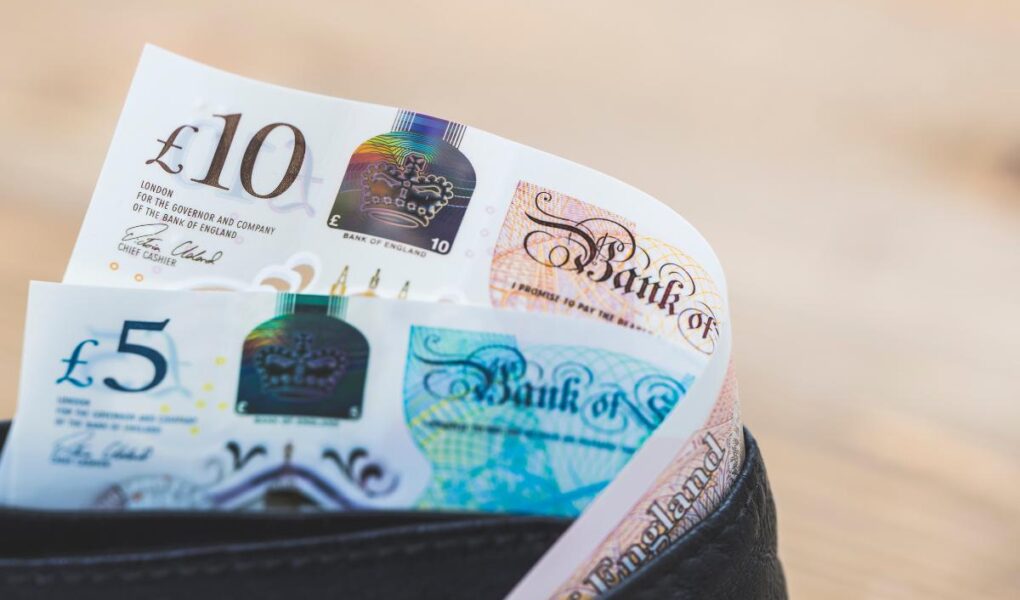 A close-up of a black wallet with British banknotes, including a £10 note and a £5 note, partially visible and sticking out. The background is softly blurred with a neutral, light brown tone.