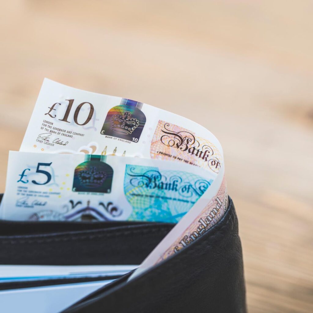 A close-up of a black wallet with British banknotes, including a £10 note and a £5 note, partially visible and sticking out. The background is softly blurred with a neutral, light brown tone.