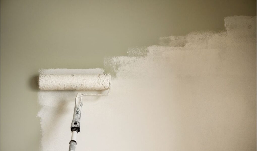 A paint roller on an extension pole is being used to apply fresh white paint over a light green wall, with part of the wall still green and the rest coated in white paint. The roller is mostly covered in paint, and visible brush strokes remain.