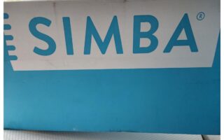 A close-up photo of a blue and white box with "SIMBA" printed in large blue letters across the side, likely packaging for a Simba Hybrid mattress. The text is centered, with indoor lighting casting partial shadows in the background.
