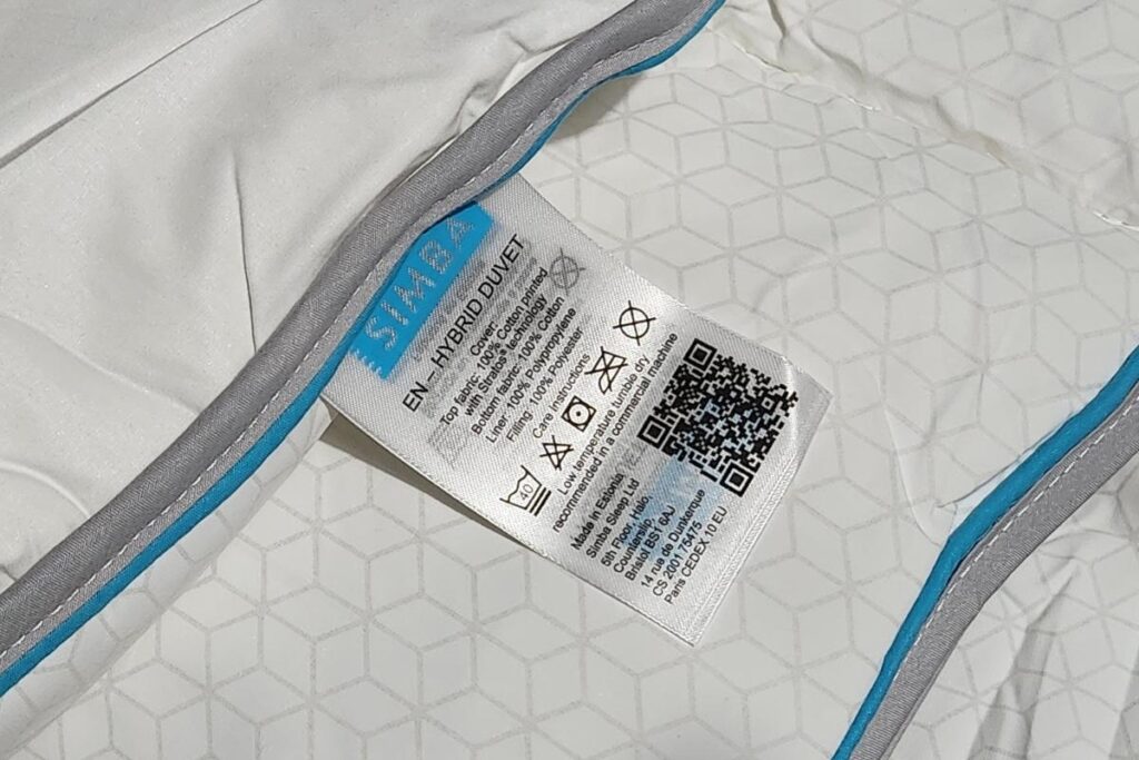 Close-up of a white duvet with a light grey and blue trim, featuring a fabric care label. The label displays IKEA branding, washing symbols, a QR code, and text in multiple languages—perfect for pairing with your Simba Hybrid mattress. Subtle geometric pattern shown.