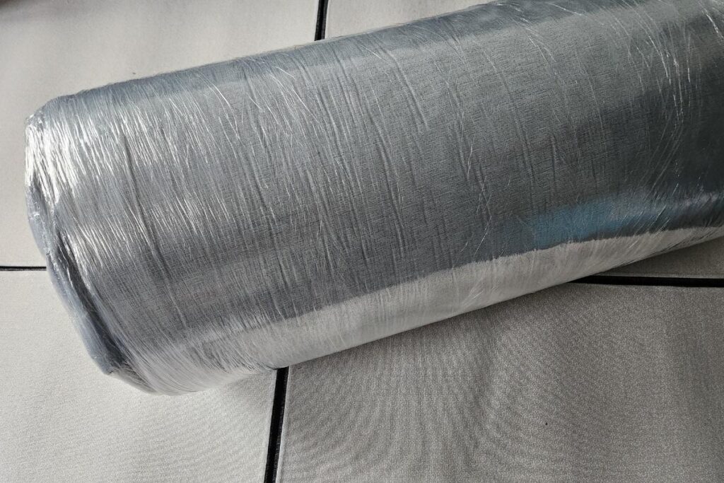 A close-up view of a large roll of silver-colored mesh, reminiscent of the fine texture found in Simba Hybrid mattresses, resting diagonally on light gray, square floor tiles with visible grout lines and a metallic sheen.