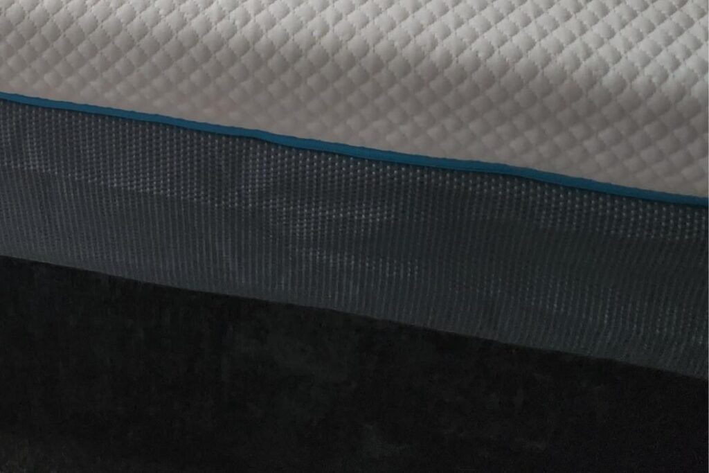A close-up view of a Simba Hybrid mattress, showing a white quilted top layer, a thin blue trim, and a dark textured fabric side panel. The image emphasizes the different materials and textures of the mattress.