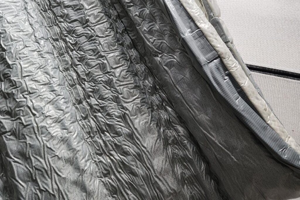 Close-up of a shiny, crinkled silver thermal blanket or heat-reflective material with quilted texture, bordered by a thick edge lined with duct tape—resembling the innovative layers found in a Simba Hybrid. A smooth, light-colored fabric is visible behind.