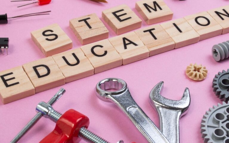 Wooden blocks spelling "STEM EDUCATION" are surrounded by various tools and objects, including wrenches, gears, a clamp, a paintbrush, wires, and electronic components, all arranged on a pink background, symbolizing science, technology, engineering, and math.