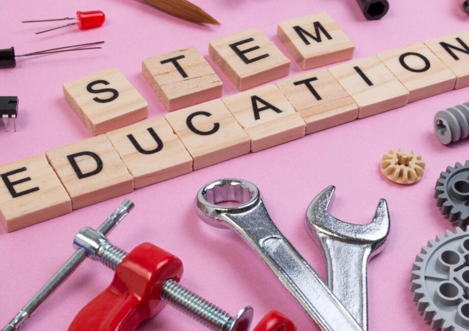 Wooden blocks spelling "STEM EDUCATION" are surrounded by various tools and objects, including wrenches, gears, a clamp, a paintbrush, wires, and electronic components, all arranged on a pink background, symbolizing science, technology, engineering, and math.