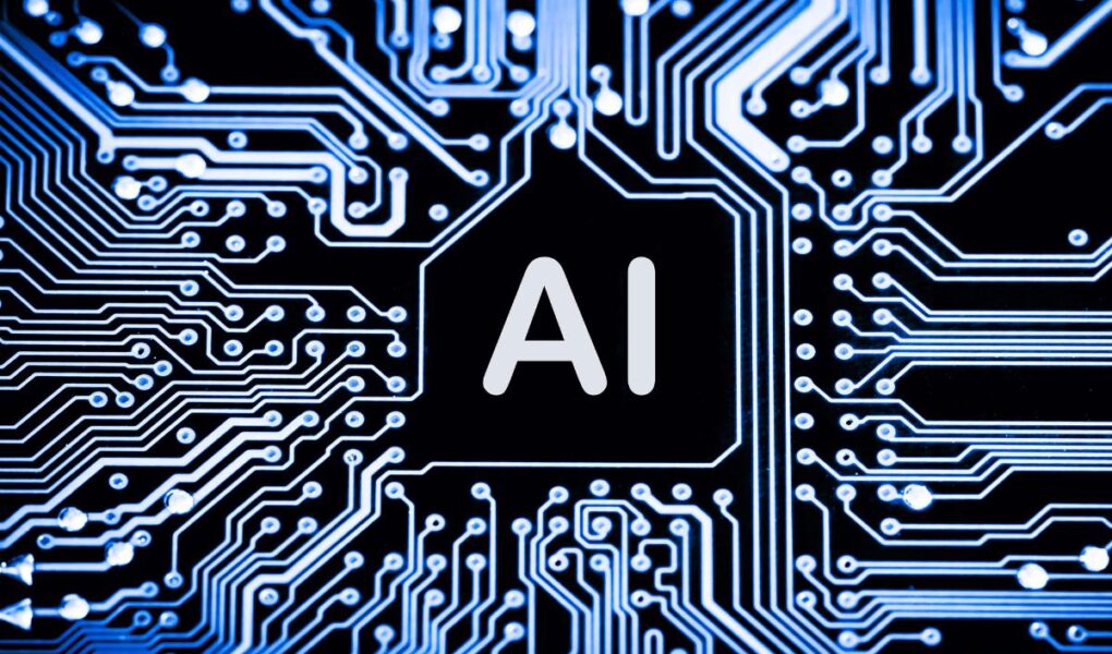 A close-up image of a printed circuit board with glowing blue lines and patterns. In the center, a black hexagon contains the large, white letters "AI," symbolizing artificial intelligence technology.