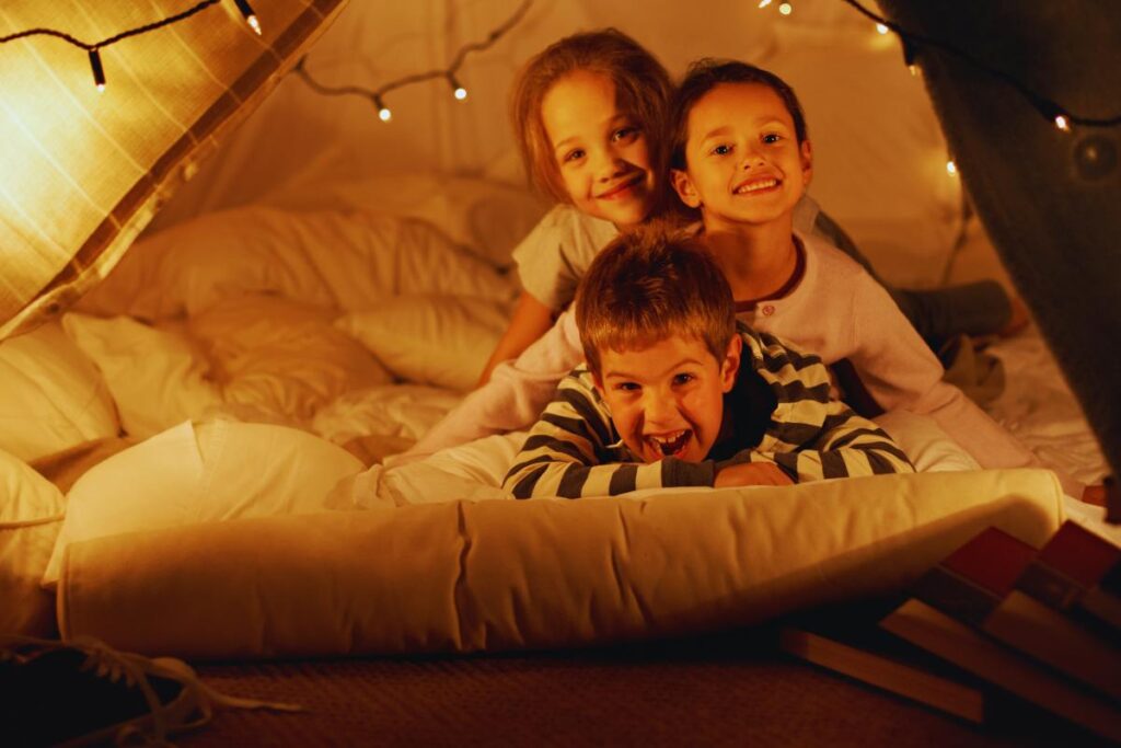 Three smiling children are piled on top of each other inside a cosy blanket fort decorated with fairy lights. Soft cushions and blankets surround them, and books are stacked nearby, creating a warm, playful indoor atmosphere.