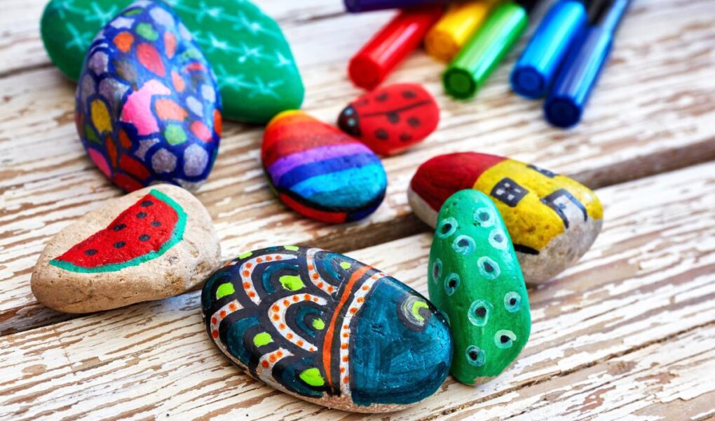 Several smooth rocks painted with colorful designs—watermelon, rainbow stripes, houses, dots, ladybugs, and abstract patterns—are scattered on a rustic wooden surface. Six markers in red, orange, yellow, green, blue, and purple lie in the background.