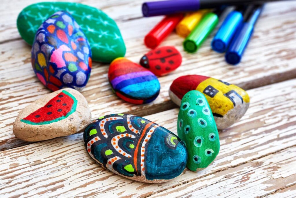 Several smooth rocks painted with colorful designs—watermelon, rainbow stripes, houses, dots, ladybugs, and abstract patterns—are scattered on a rustic wooden surface. Six markers in red, orange, yellow, green, blue, and purple lie in the background.