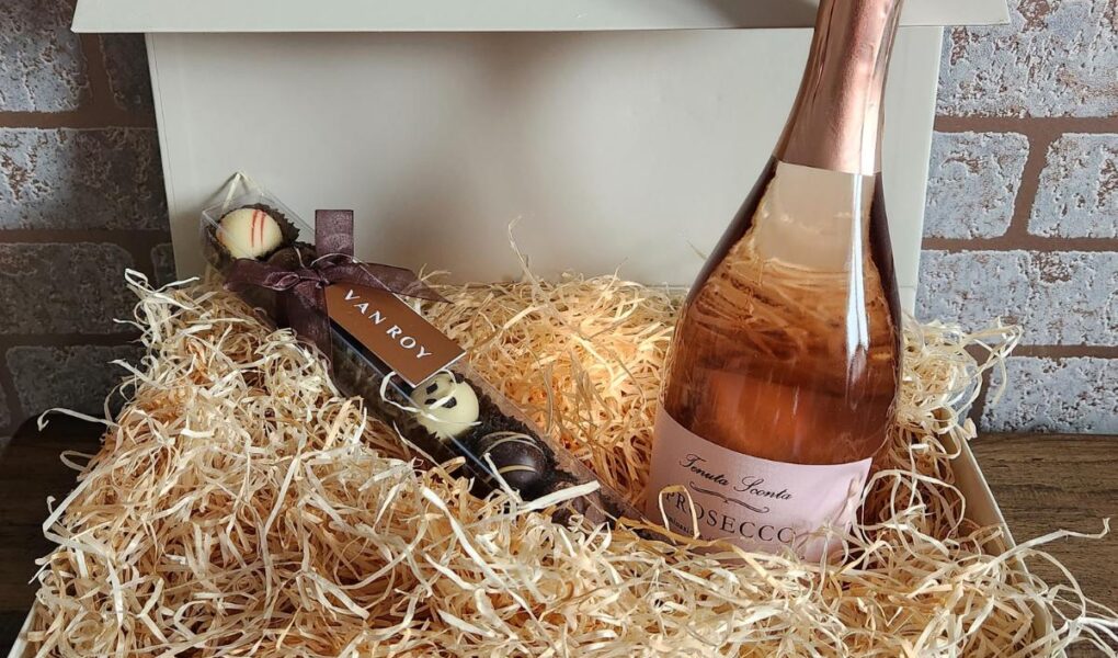 A white gift box filled with straw contains a bottle of rose prosecco and a stick of assorted chocolates. The box is open, sitting on a wooden surface against a brick wall background.