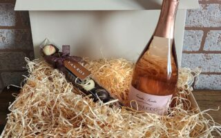 A white gift box filled with straw contains a bottle of rose prosecco and a stick of assorted chocolates. The box is open, sitting on a wooden surface against a brick wall background.