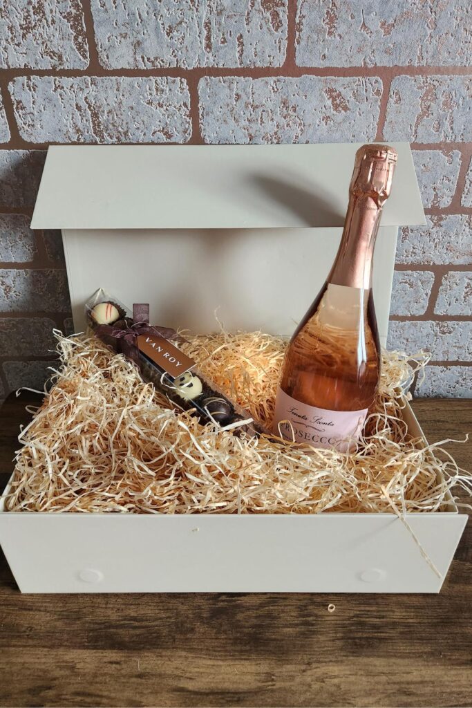 A white gift box filled with straw contains a bottle of rose prosecco and a stick of assorted chocolates. The box is open, sitting on a wooden surface against a brick wall background.