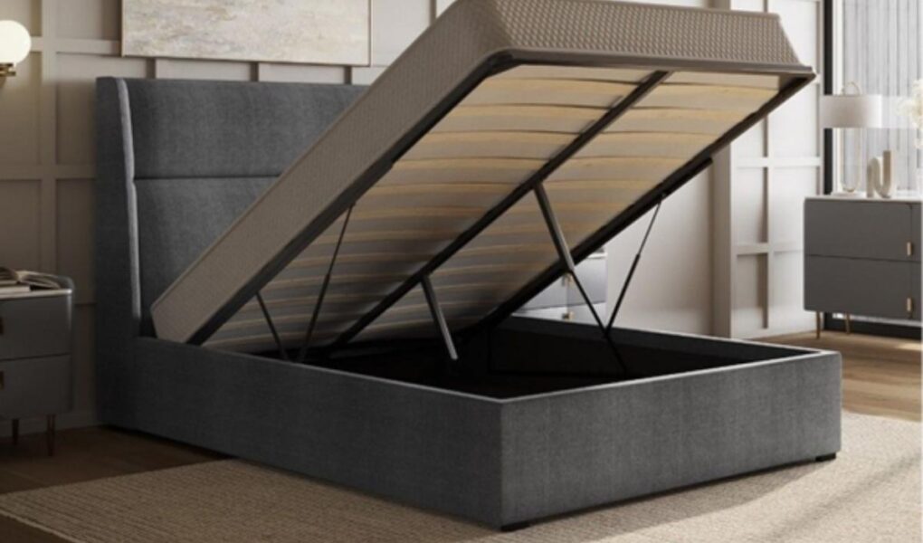 A modern upholstered storage bed with the mattress platform lifted, revealing a spacious storage compartment underneath. The bed is dark gray, with a tall headboard, in a stylish bedroom with neutral decor and wooden flooring.