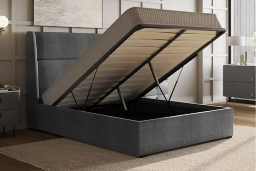 A modern upholstered storage bed with the mattress platform lifted, revealing a spacious storage compartment underneath. The bed is dark gray, with a tall headboard, in a stylish bedroom with neutral decor and wooden flooring.