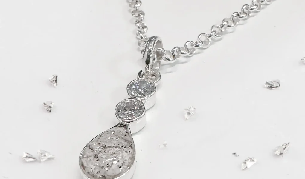 A silver necklace with a pendant featuring two round clear stones above a teardrop-shaped stone with a speckled, gray-white appearance, displayed on a white surface with small scattered silver fragments.