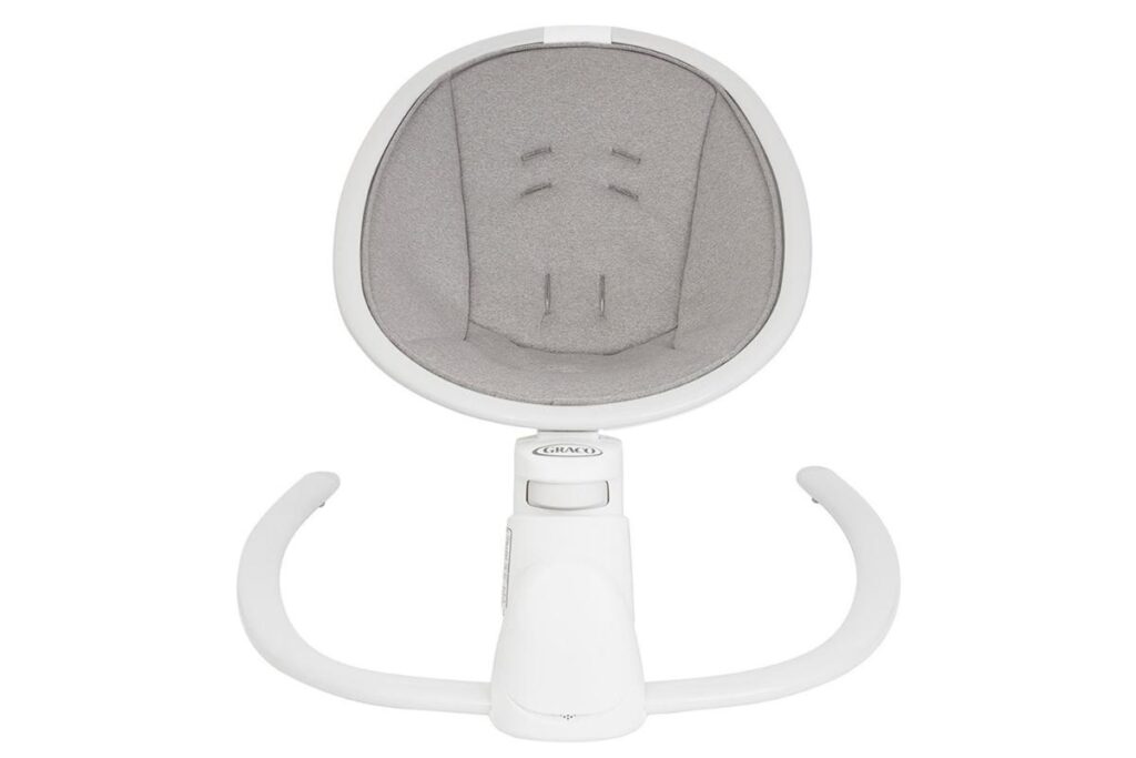 A top-down view of a modern baby rocker seat with a white frame and light gray padded fabric inside. The seat has built-in harness slots and two curved white legs extending forward, designed for stability and gentle rocking.