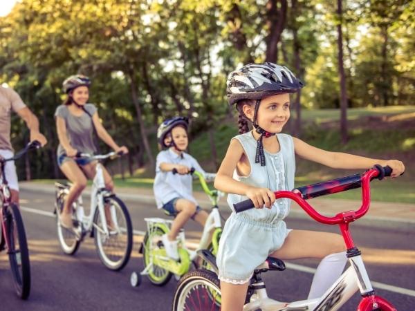 Four children ride bicycles on a sunlit, tree-lined road. They all wear helmets and casual summer clothing. The lead girl smiles confidently; behind her, a younger child rides a bike with training wheels. Two more kids follow, each focused on riding.