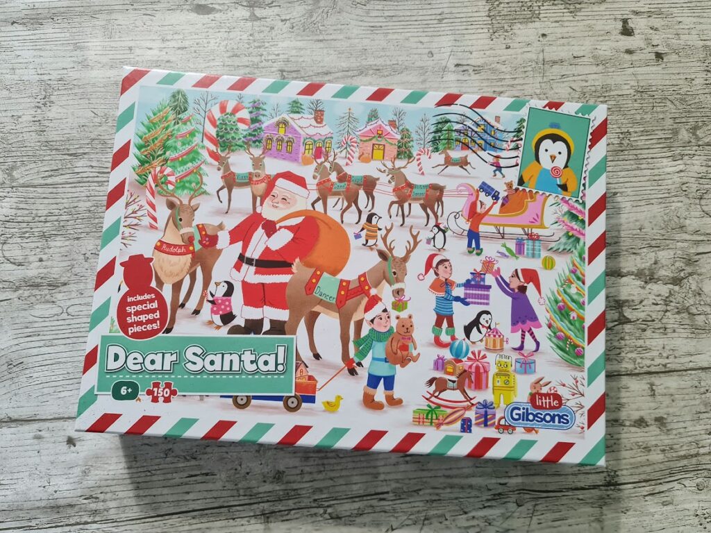 A colorful puzzle box titled "Dear Santa!" shows Santa with his reindeer, elves, and children in a snowy village. Presents, sleigh, decorated trees, and festive houses are in the background. The box features a penguin stamp and striped border. For ages 6+.