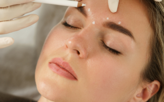 A woman with closed eyes lies back as a gloved hand marks dots above her eyebrow with a white pencil, possibly preparing for a cosmetic procedure. Her expression is calm and relaxed.