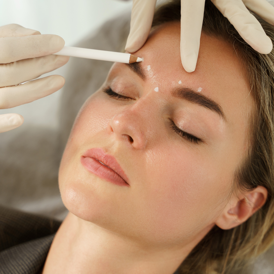A woman with closed eyes lies back as a gloved hand marks dots above her eyebrow with a white pencil, possibly preparing for a cosmetic procedure. Her expression is calm and relaxed.