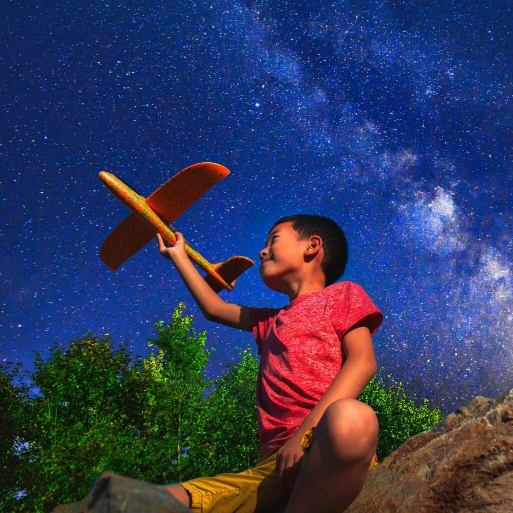 A young boy in a red shirt and yellow shorts sits outdoors at night, holding a toy orange airplane aloft. Behind him, the sky is filled with bright stars and the Milky Way. Green trees and rocky ground are visible in the foreground.