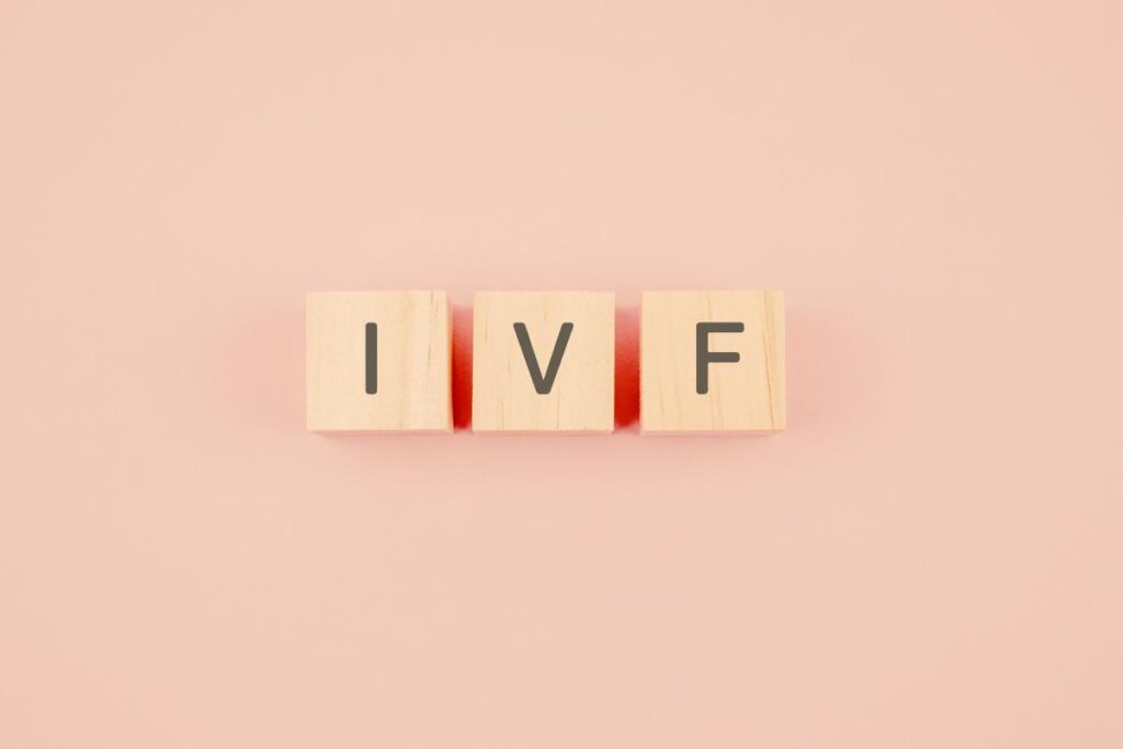 Three wooden blocks, each with a single black letter on the front, spell out "IVF" against a soft pink background. The blocks are evenly spaced and aligned in a straight horizontal row.