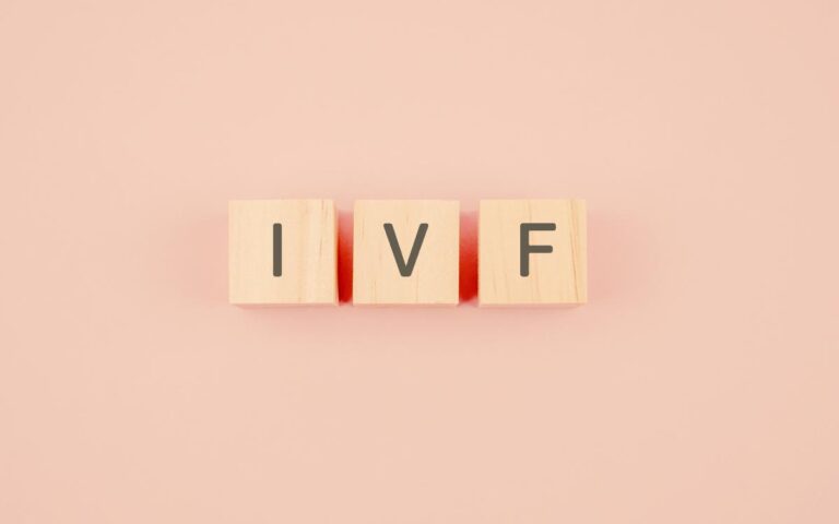 Three wooden blocks, each with a single black letter on the front, spell out "IVF" against a soft pink background. The blocks are evenly spaced and aligned in a straight horizontal row.